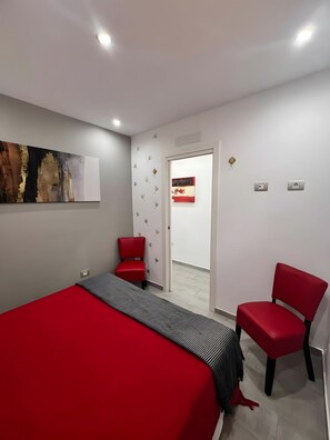 Ein Hotelzimmer mit einem Bett, zwei roten Stühlen, einer roten und schwarzen Bettdecke, einem Wandbild und einer Tür zu einem anderen Raum.