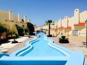 Ein Poolbereich mit einer geschwungenen Wasseranlage, Palmen und Gebäuden mit roten Ziegeldächern.