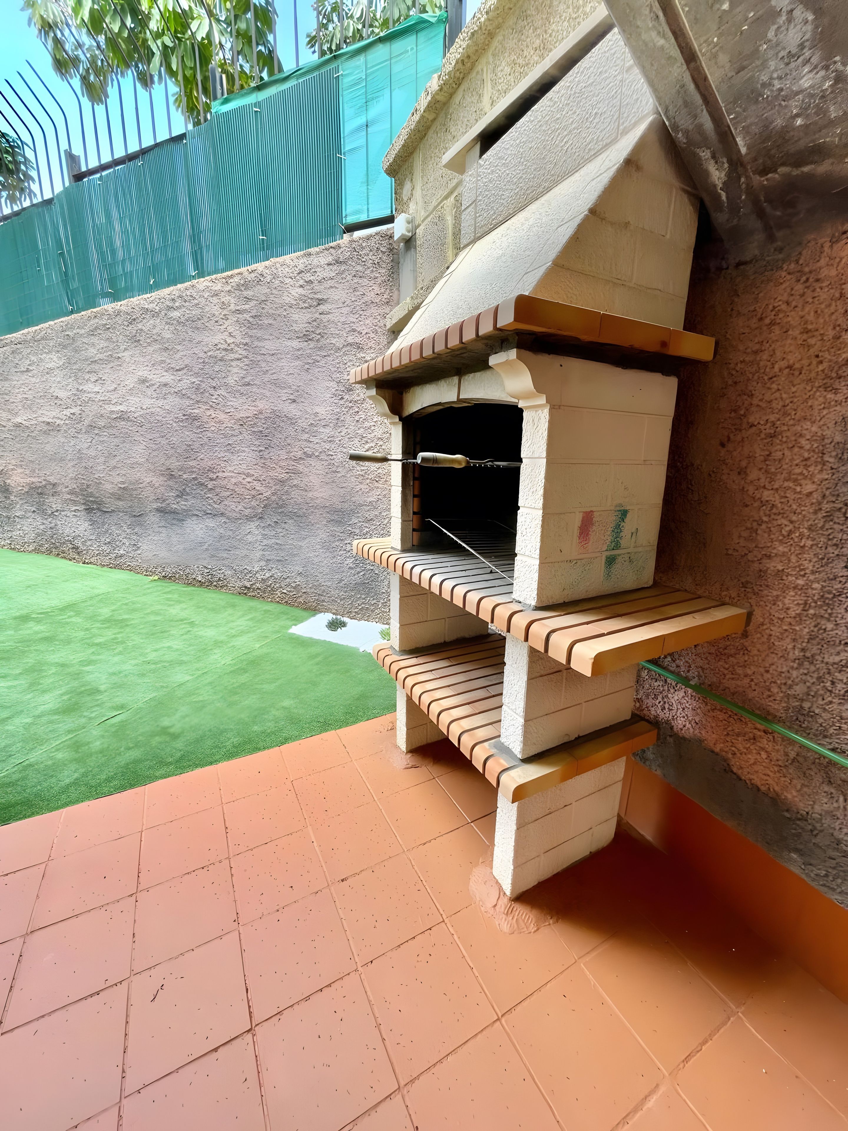 Een buitenoven gebouwd van bakstenen, met een plank, gelegen voor een muur met een groen gazon en een hek op de achtergrond.