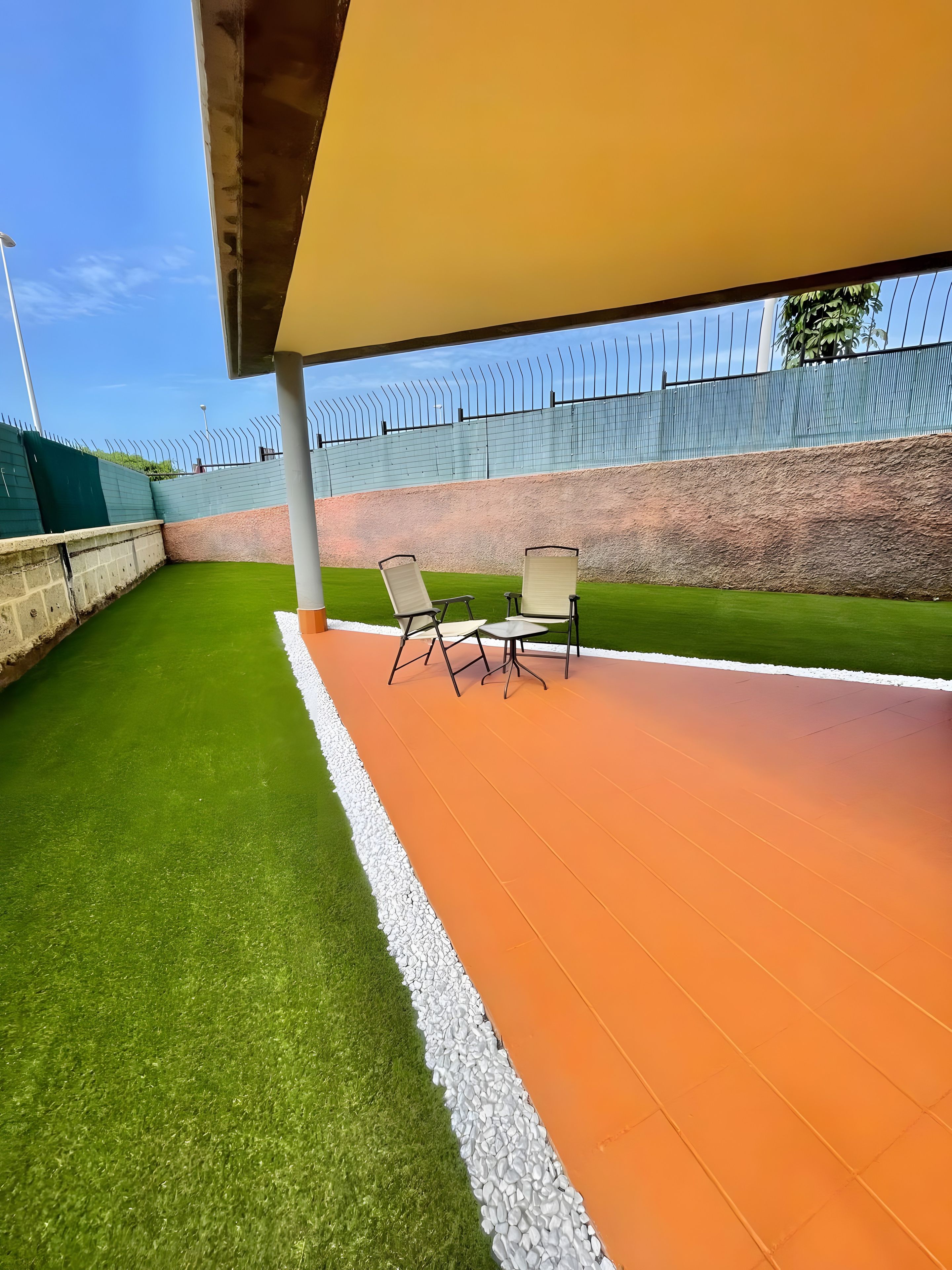 An outdoor seating area with artificial grass and a paved walkway, surrounded by a high fence.
