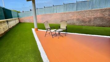 An outdoor seating area with artificial grass and a paved walkway, surrounded by a high fence.