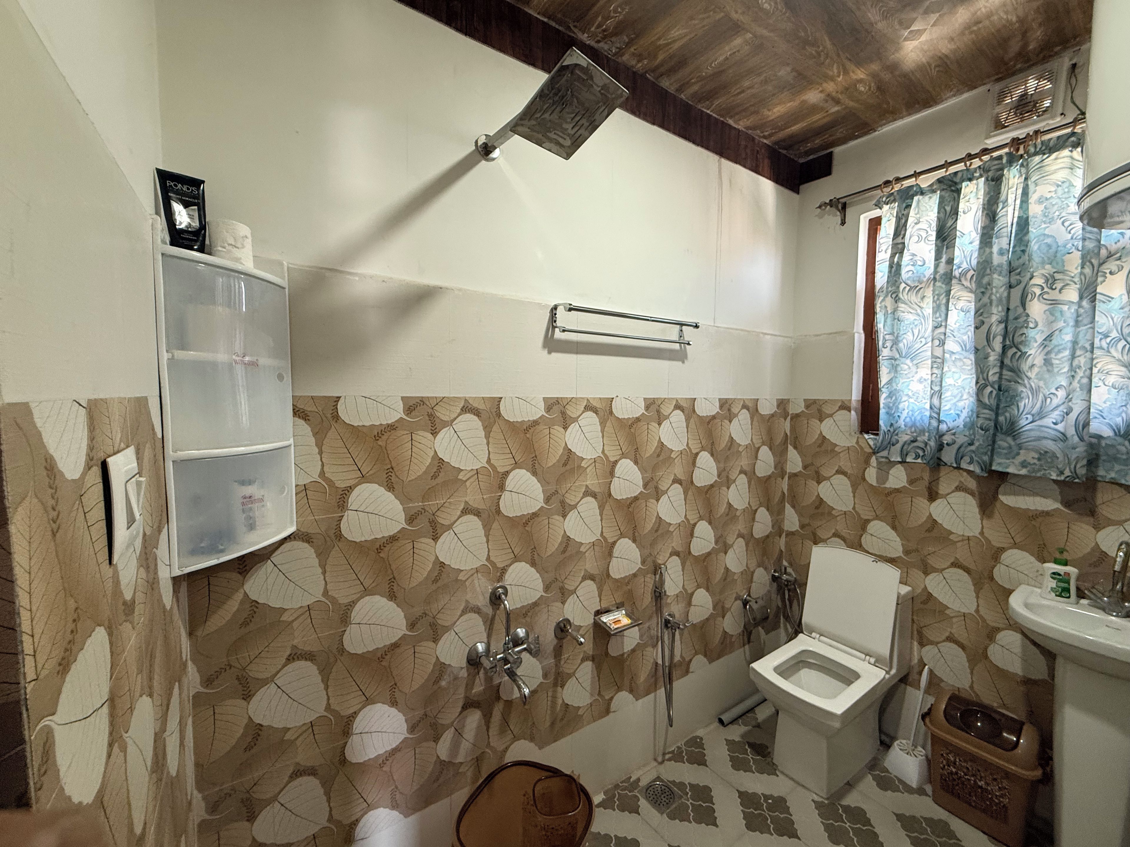 Casa de banho com chuveiro, sanita e lavatório. Inclui um porta-toalhas e uma janela com cortinas.