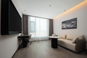 Een moderne hotelkamer met een flatscreen televisie, een bank, een klein rond tafeltje en een groot raam met gordijnen.