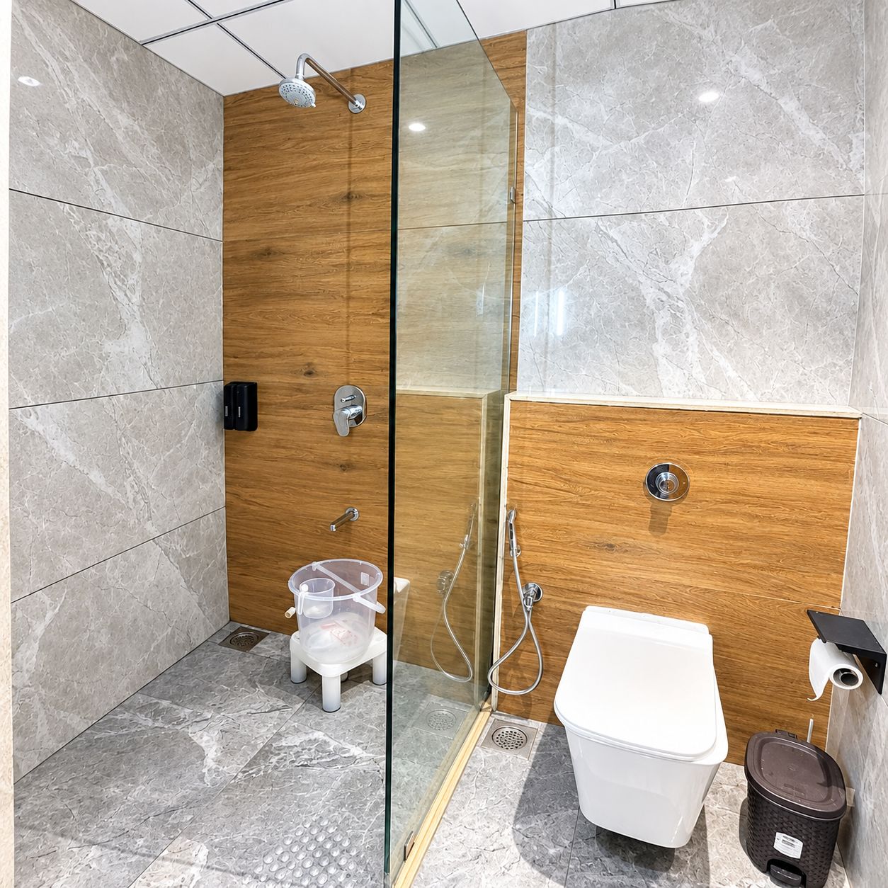 A modern bathroom with a glass-enclosed shower, a wooden vanity, a toilet, and a trash bin.