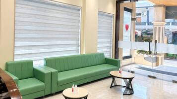 Un área de recepción con un sofá verde, dos mesas redondas y una puerta de vidrio que da al exterior.