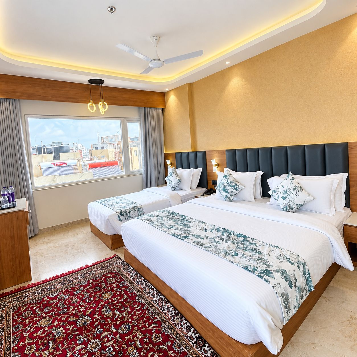 A hotel room with two beds, a large window, a ceiling fan, and a red patterned rug.