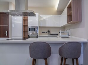 Une cuisine moderne avec des meubles blancs, une hotte de cuisinière en acier inoxydable, un four et un évier. On trouve deux tabourets de bar gris devant le comptoir.