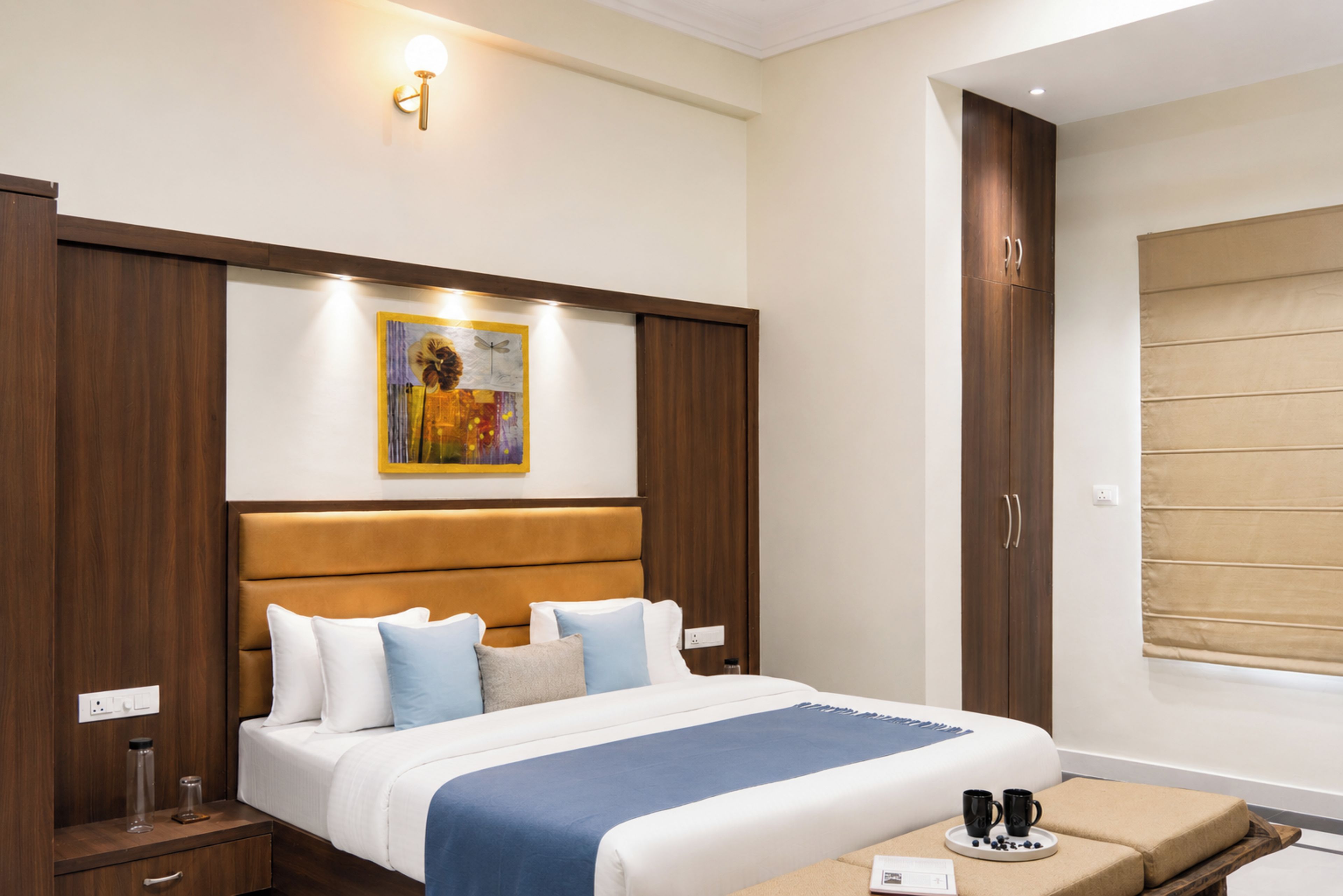 A hotel room with a bed, bedside tables, a nightstand, a wall-mounted artwork, a window with blinds, and a wardrobe.