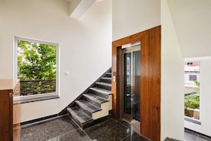 A staircase with a wooden handrail, a door with a glass panel, and a window with a view of greenery. - Alaya Stays Riva Amar Villa 3BHK (Udaipur)
