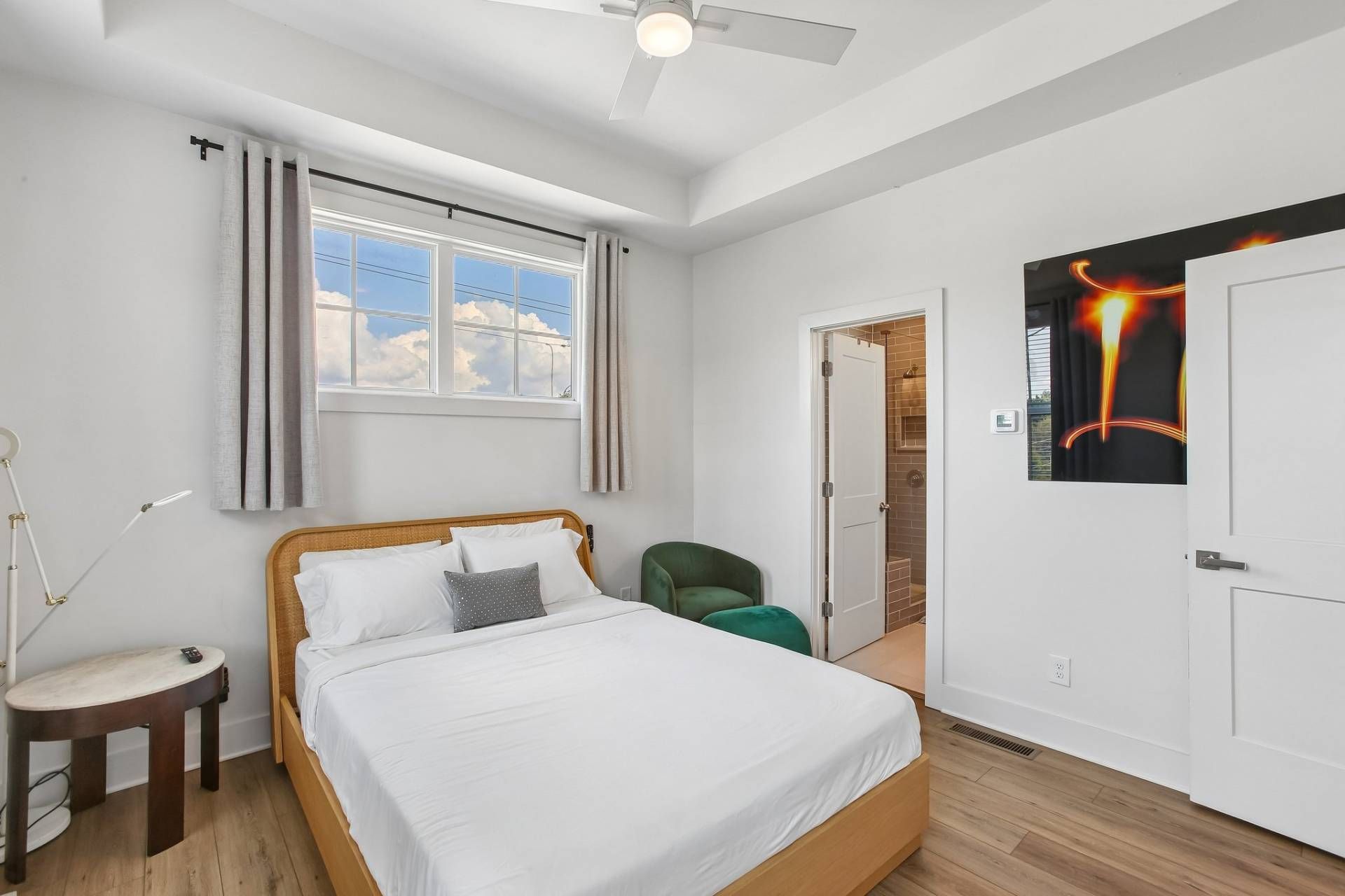 Une chambre à coucher comprenant un lit, une chaise, une table, une fenêtre avec des rideaux, un ventilateur de plafond et une porte ornée d’un tableau.