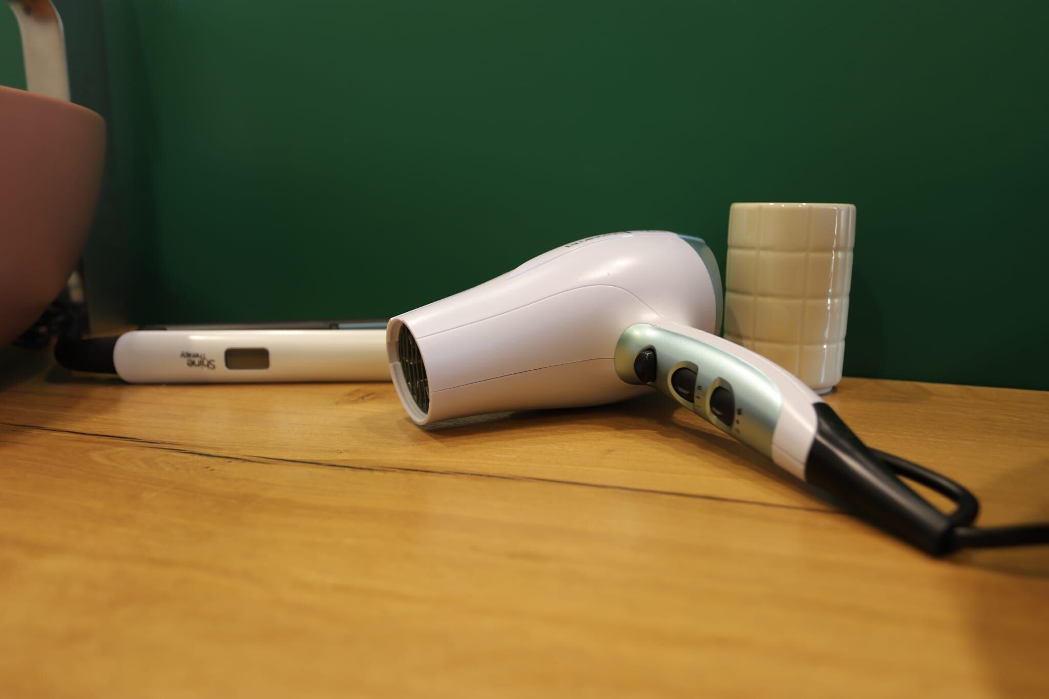 A hair dryer and a cup on a wooden surface against a green wall.