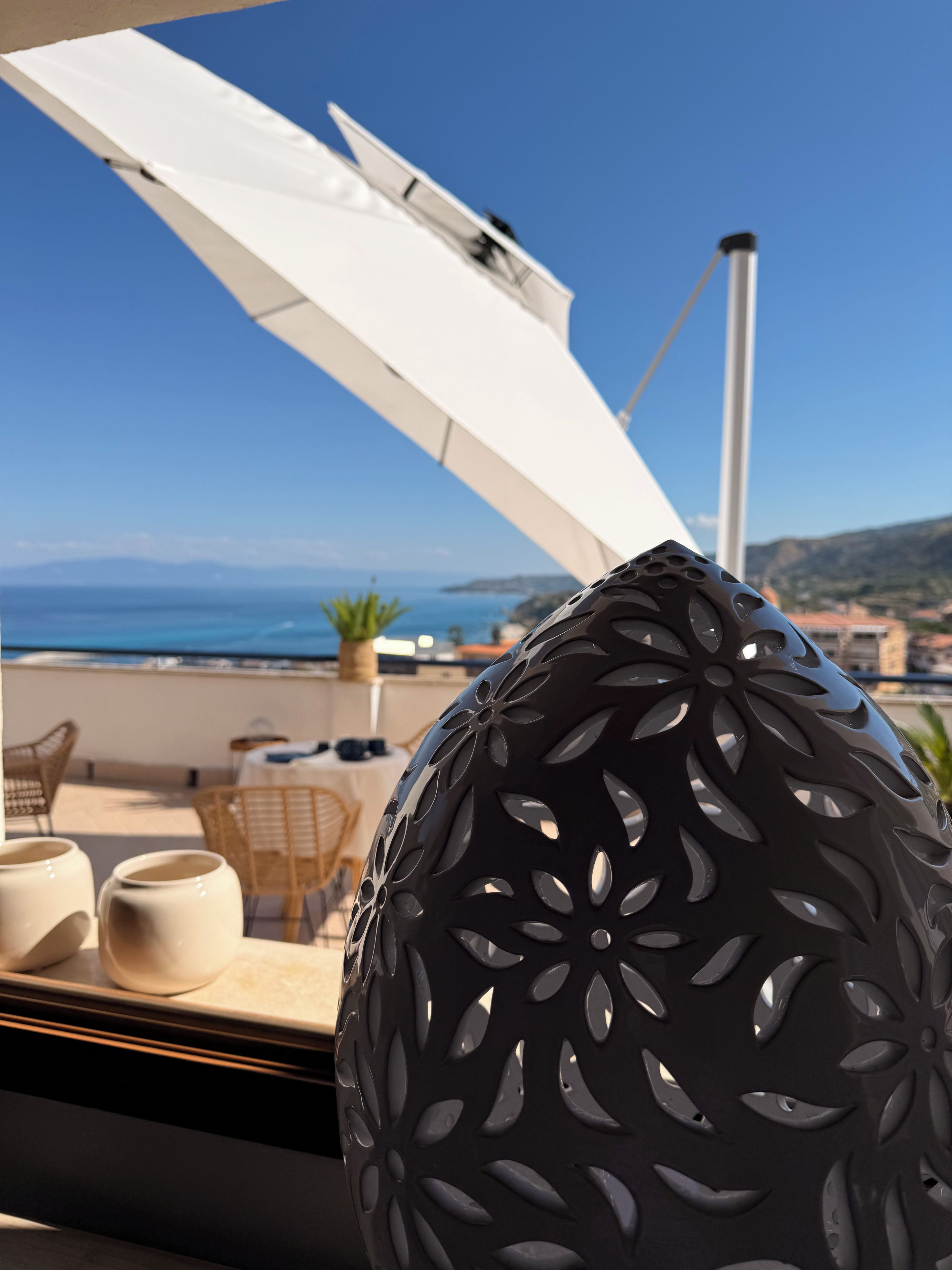 A decorative black vase with a floral pattern on a table, overlooking a coastal view with a clear sky.