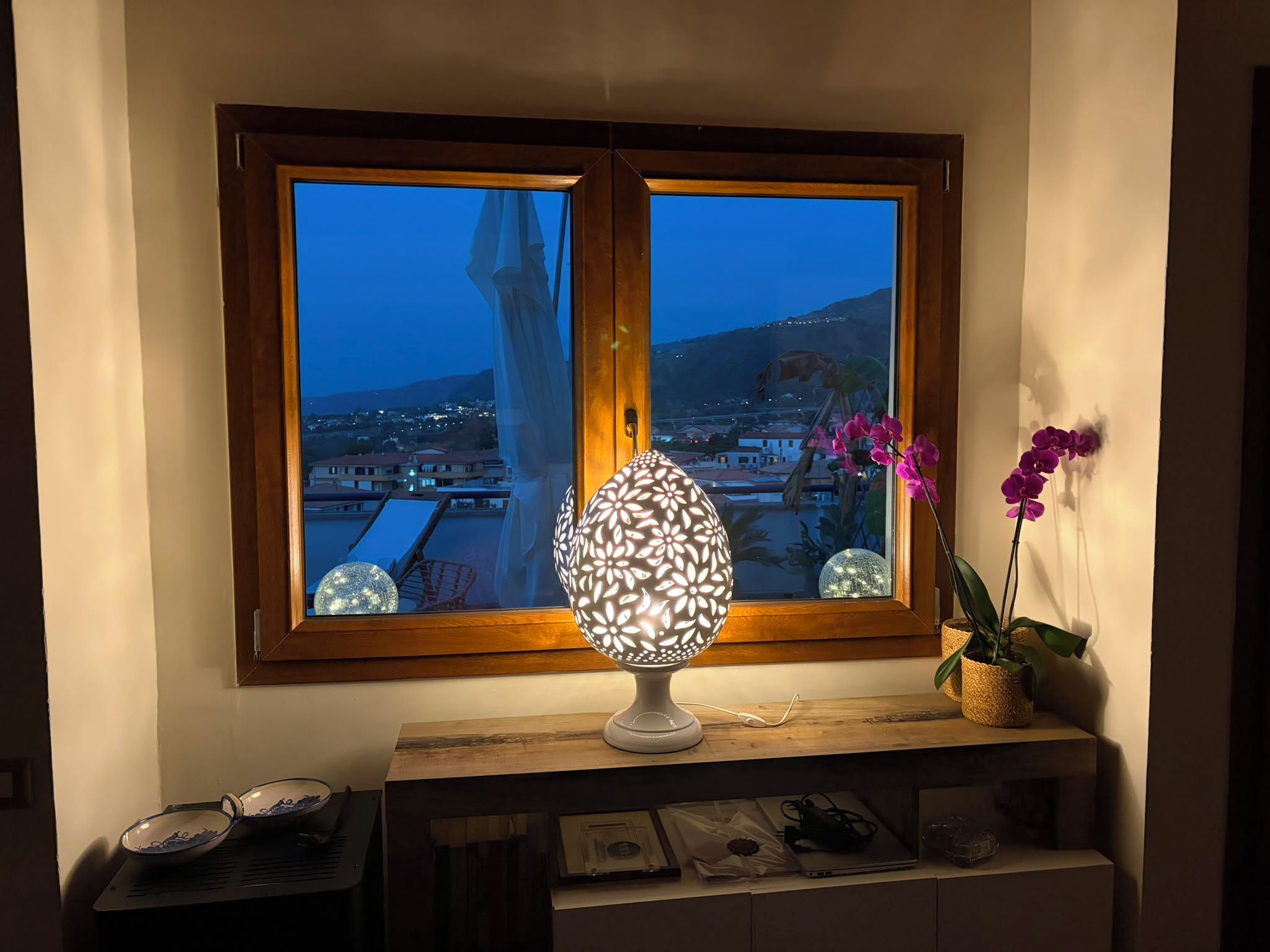 Ein Zimmer mit Fensterblick auf eine Stadtlandschaft im Abendrot, einer dekorativen Lampe und einer Orchideenpflanze im Blumentopf.