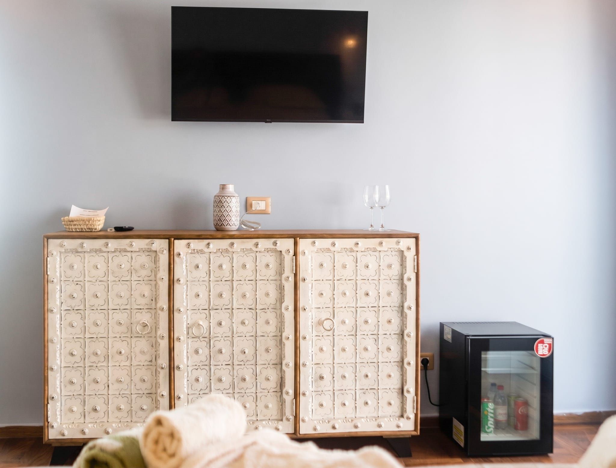 A room with a television mounted on the wall, a cabinet with decorative panels, and a refrigerator with beverages.