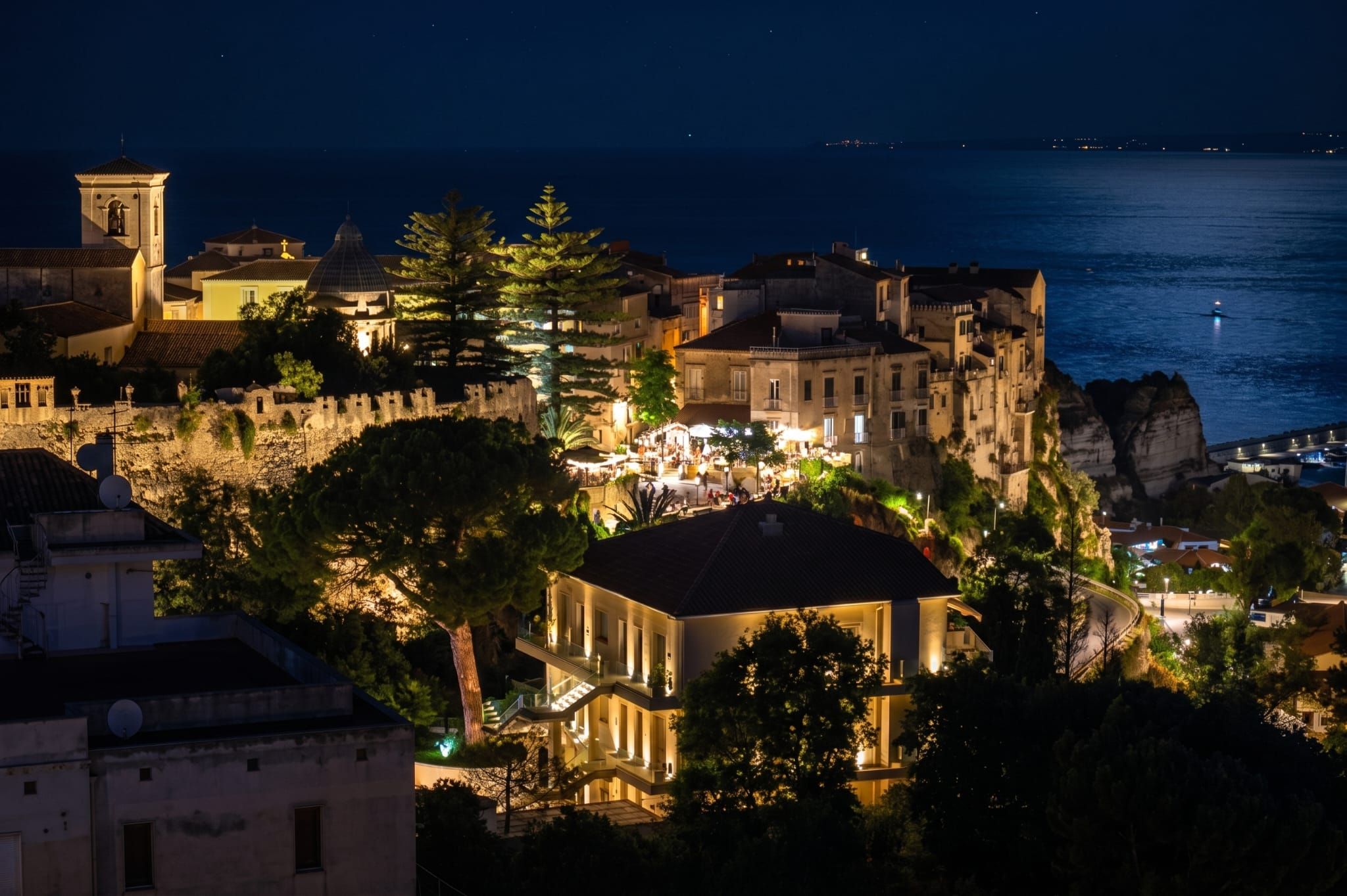 A coastal town at night with illuminated buildings, a prominent church tower, and a rocky coastline.