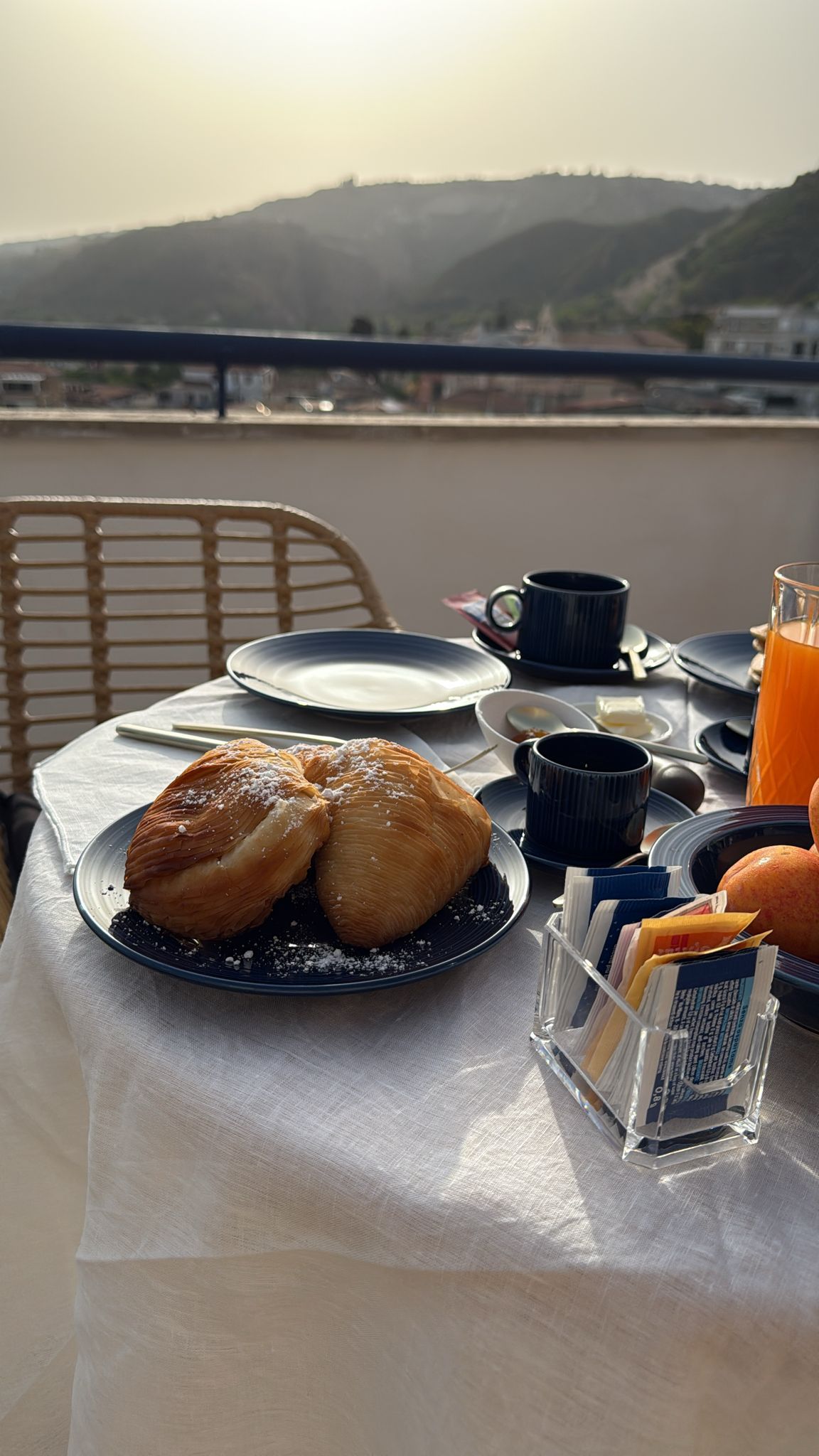 A breakfast setup with croissants on a plate, a coffee cup, a glass of orange juice, and a book holder on a table.