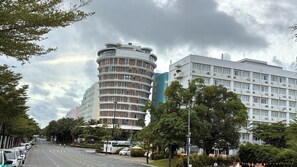Ein mehrstöckiges Gebäude mit einer geschwungenen Fassade, ein daran angrenzendes Gebäude mit Flachdach sowie eine Straße mit parkenden Autos und Bäumen.