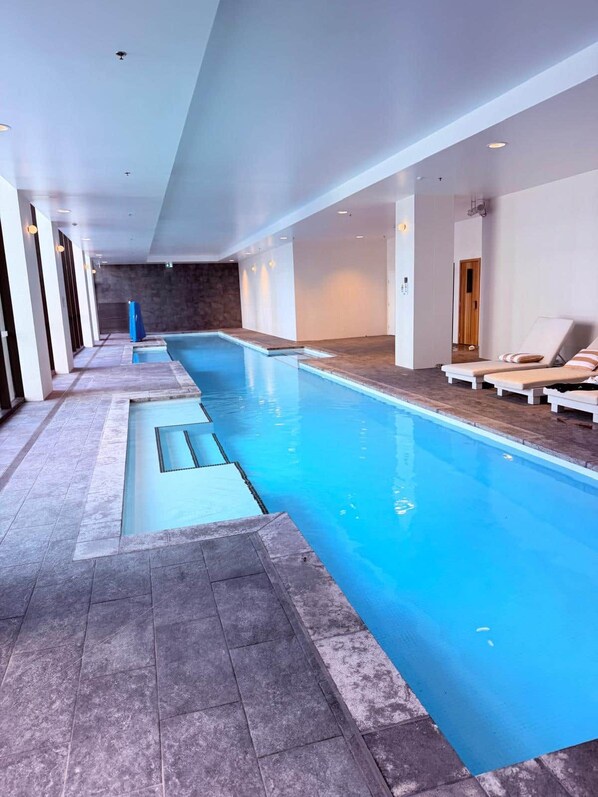 Ein Hallenbad mit klarem Wasser, gefliestem Boden und Liegestühlen an den Seiten.