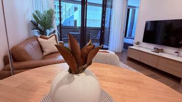 Een moderne woonkamer met een ronde houten tafel, een witte vaas met bruine bladeren, een leren bank in bruin, een televisie en een groot raam met gordijnen.