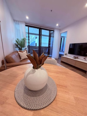 Ein modernes Wohnzimmer mit einem runden Holztisch, einer weißen Vase mit braunen Blättern, einem braunen Ledersofa, einem Fernseher und einem großen Fenster mit Vorhängen.