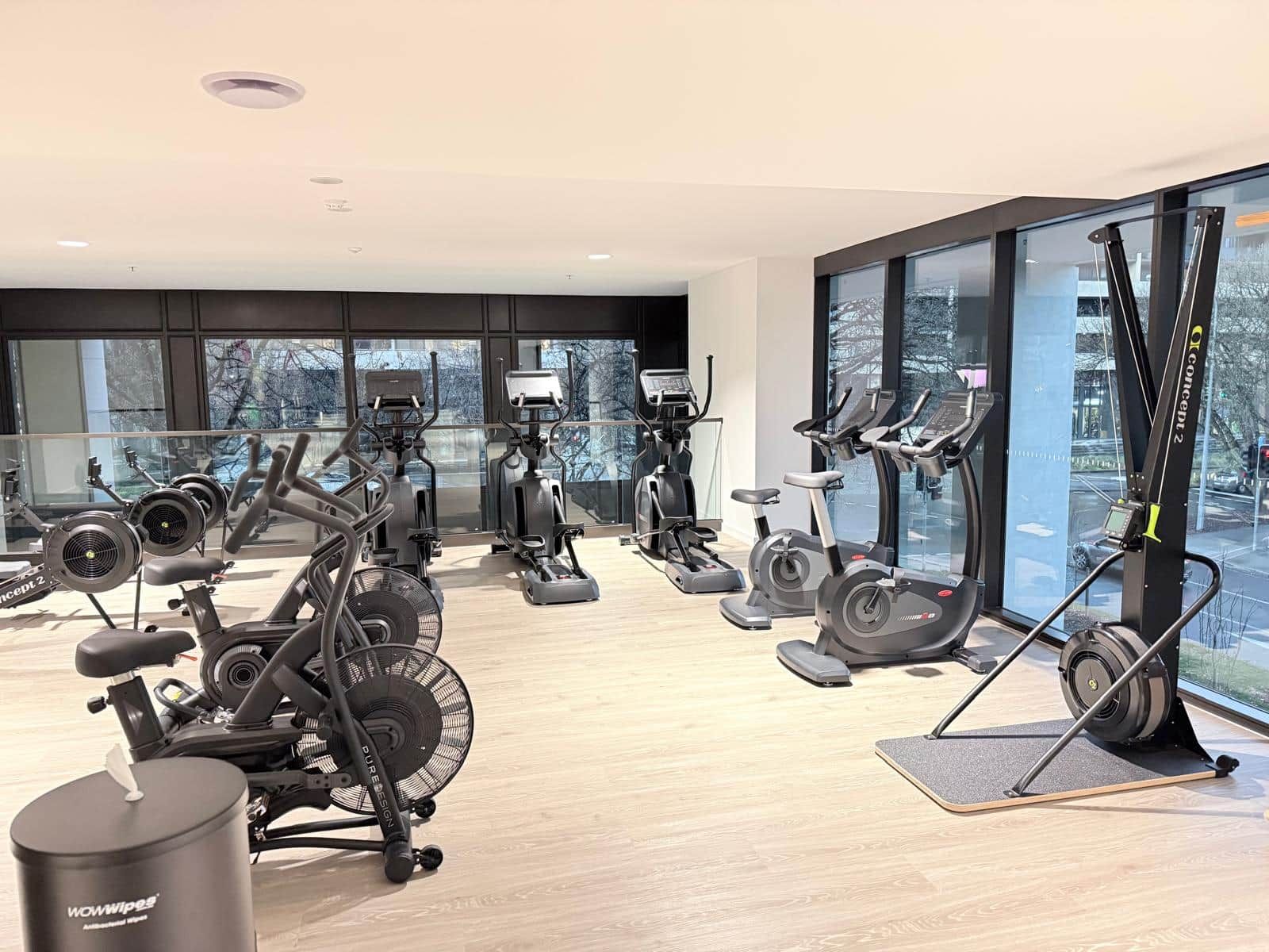 Academia moderna com diversas máquinas de cardio, uma janela ampla com vista para a cidade e uma lixeira identificada como 'WOWWipes'.