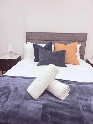 Ein ordentlich bezogenes Bett mit grauem Kopfteil, weißen Laken und mehreren Kissen in verschiedenen Farben.