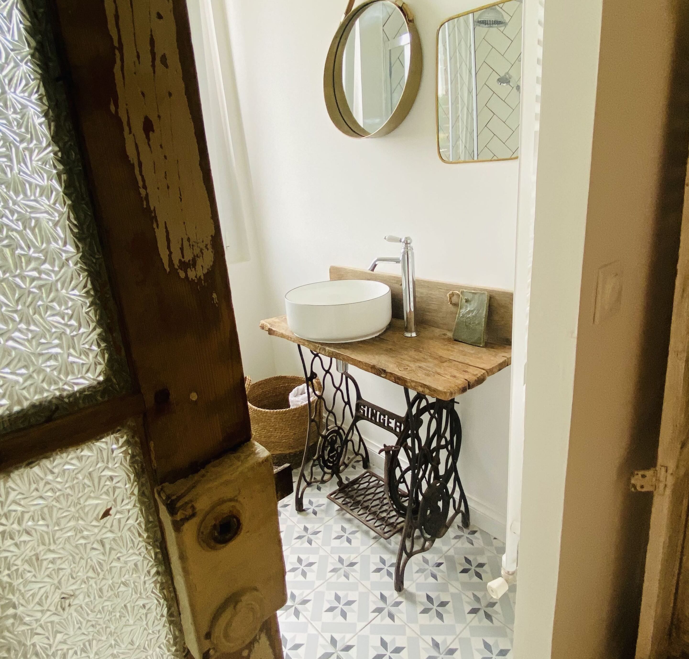 A bathroom with a vintage sewing machine sink, a round mirror, and a herringbone patterned floor.