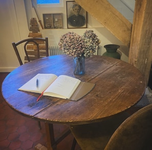 A round wooden table with a book and a pen, a vase with dried flowers, and a framed portrait on the wall.