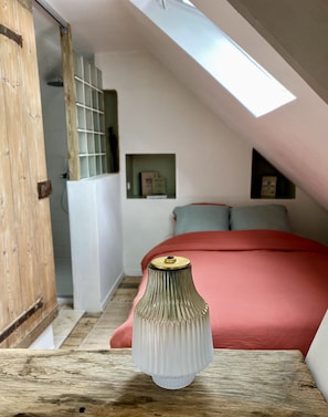 A bedroom with a sloped ceiling, a wooden door, a bedside table with a vase, and a red bedspread.