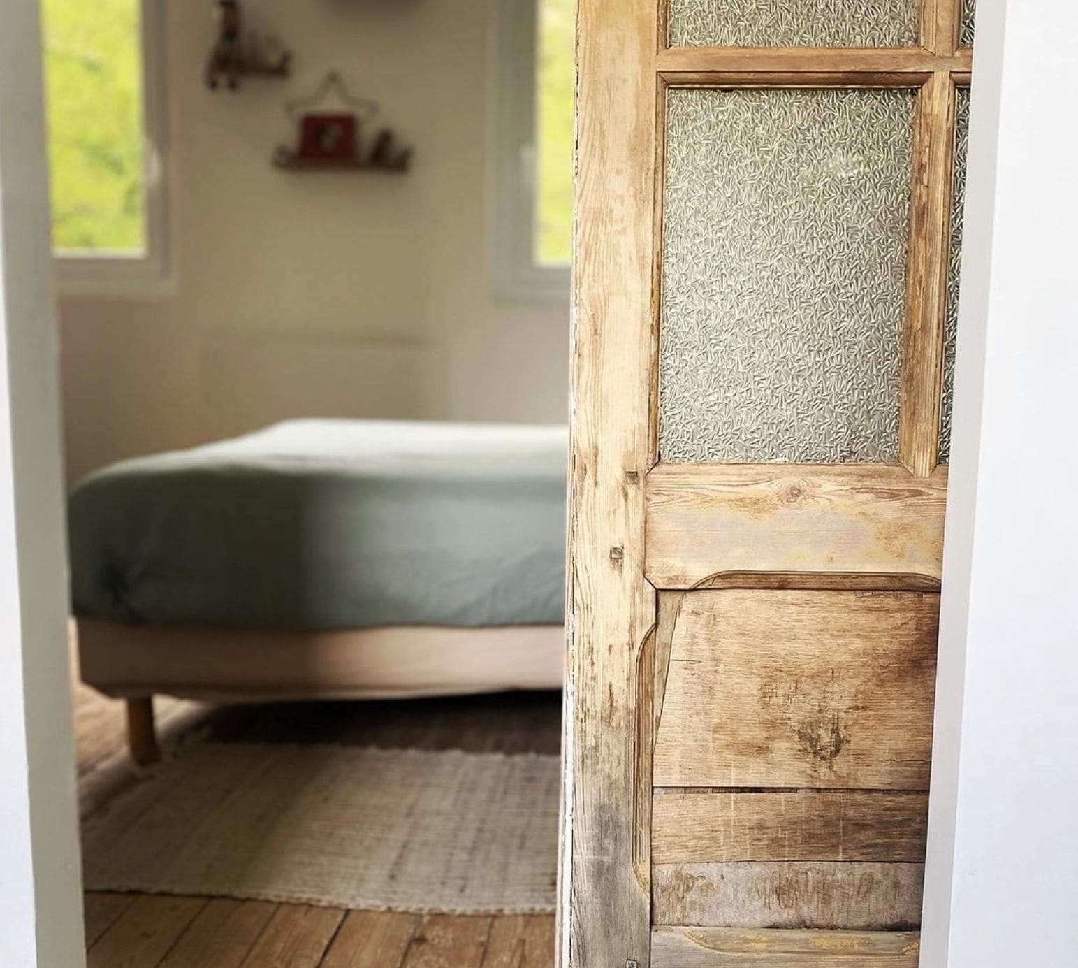 Une pièce comprenant un lit, une porte en bois avec un panneau texturé et une fenêtre donnant sur un paysage verdoyant.