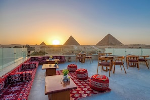 Terraza en la azotea con asientos y mesas, con vistas a las pirámides al atardecer.