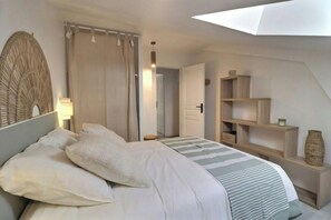 A bedroom with a bed, a bedside lamp, a woven wall headboard, a striped bedspread, a wooden shelf with a vase, and a door leading to another room. - Renovated stone house near Jaujac, Ardèche (Jaujac)