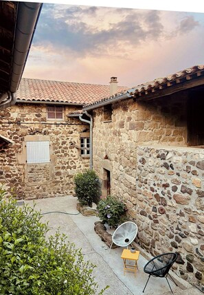 A stone building with a tiled roof, a small window, and a door. There is a chair and a table outside. - Renovated stone house near Jaujac, Ardèche (Jaujac)