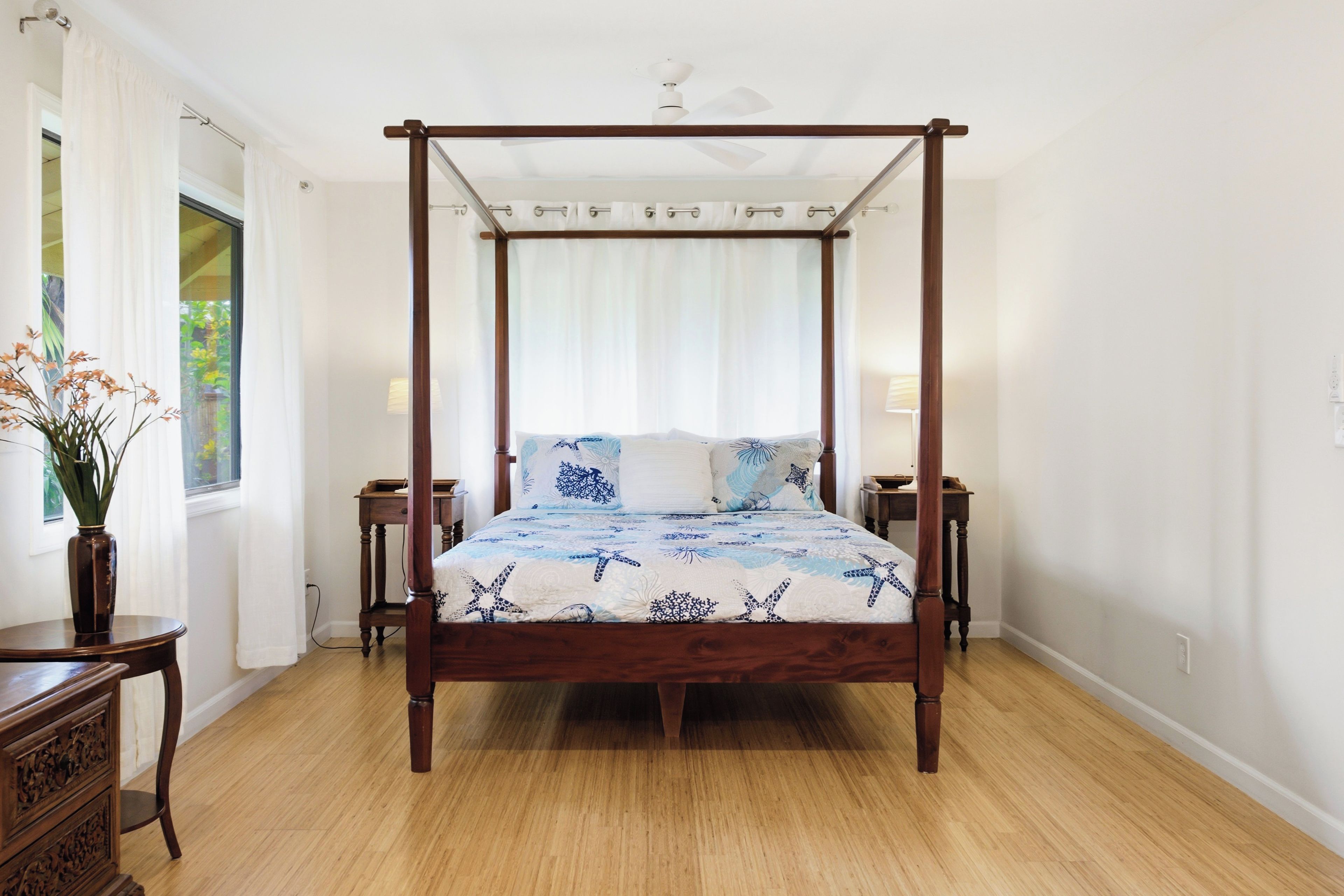 A four-poster bed with a nautical-themed bedspread, a wooden nightstand on each side, and a vase with flowers on the left side.