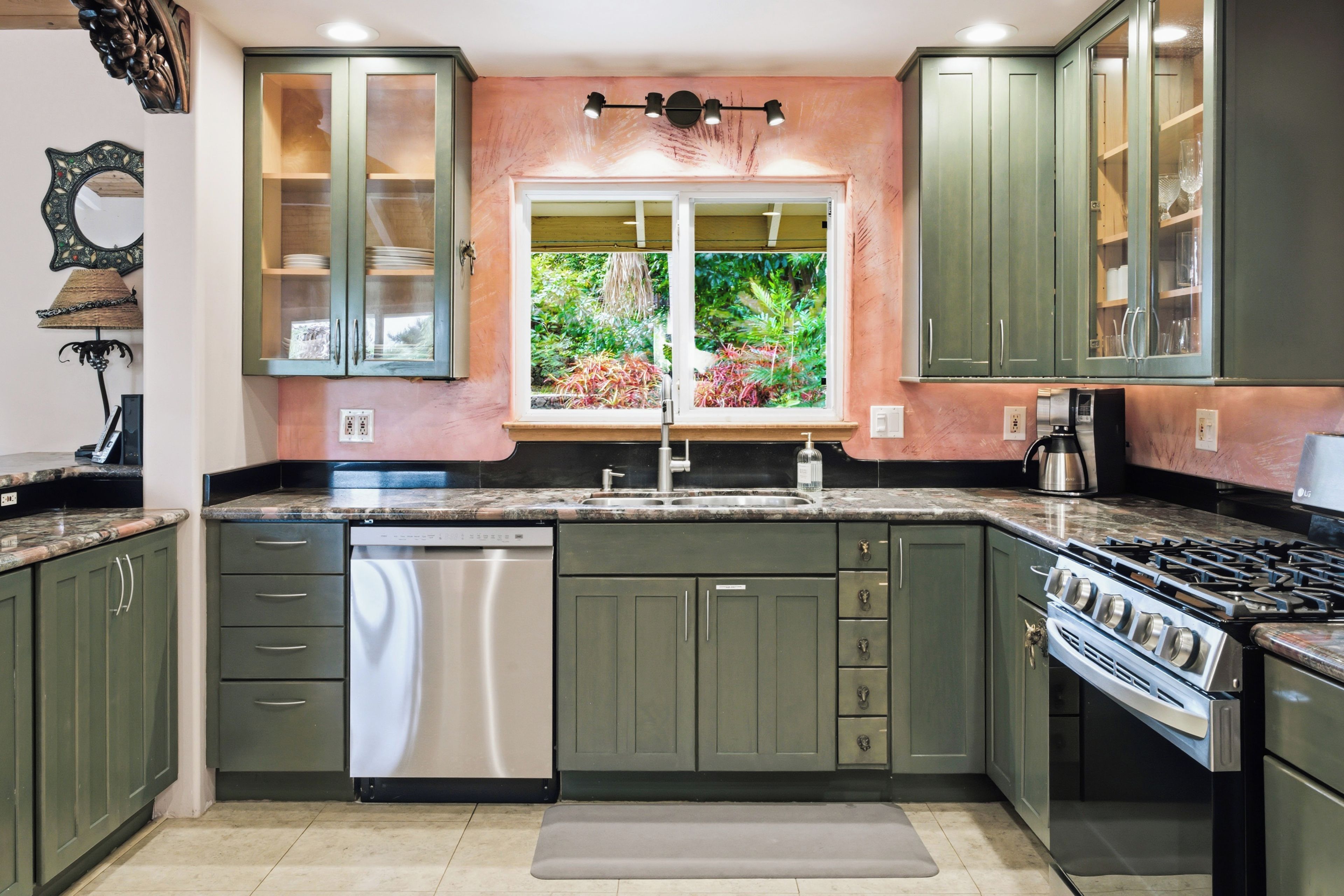 A kitchen with green cabinets, a stainless steel dishwasher, a window with a view of greenery, and a gas stove.