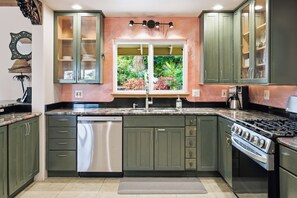 A kitchen with green cabinets, a stainless steel dishwasher, a window with a view of greenery, and a gas stove.