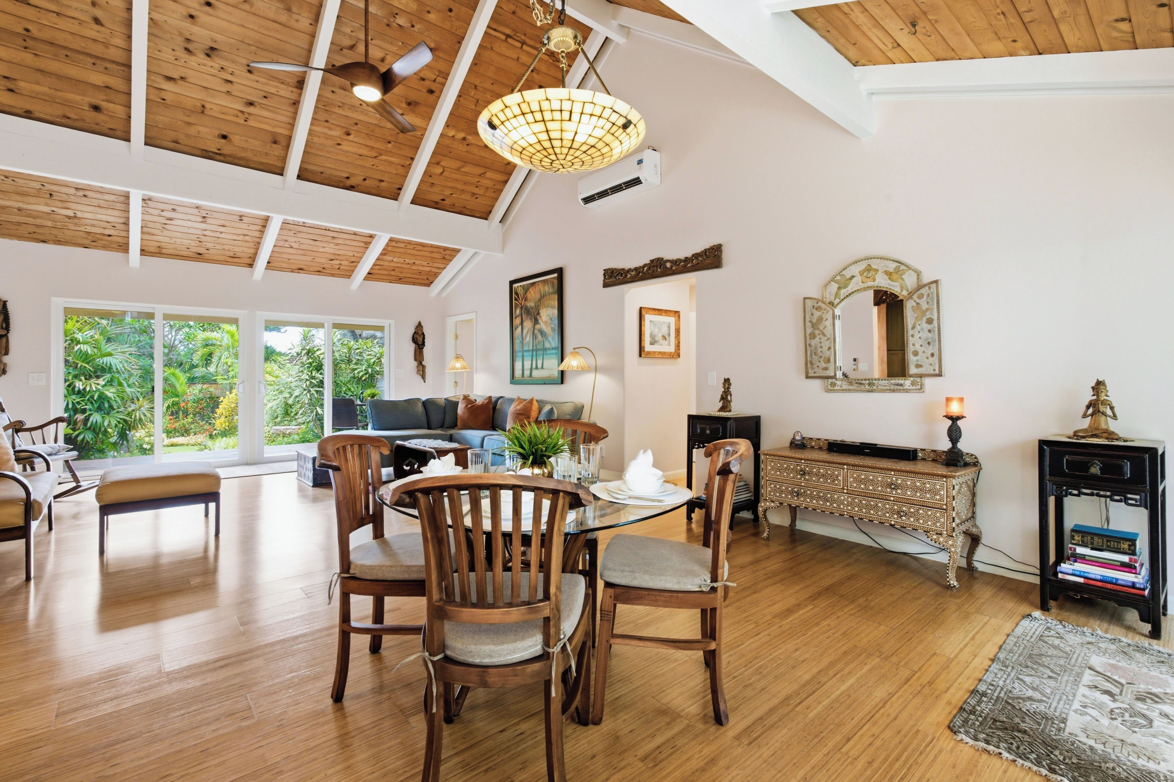 A spacious living area with a dining table, chairs, and a ceiling fan. There is a large window providing a view of the outdoors.