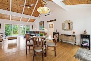 A spacious living area with a dining table, chairs, and a ceiling fan. There is a large window providing a view of the outdoors. - Lush Tropical Home Close to the Ocean - Mana Hale (Kihei)