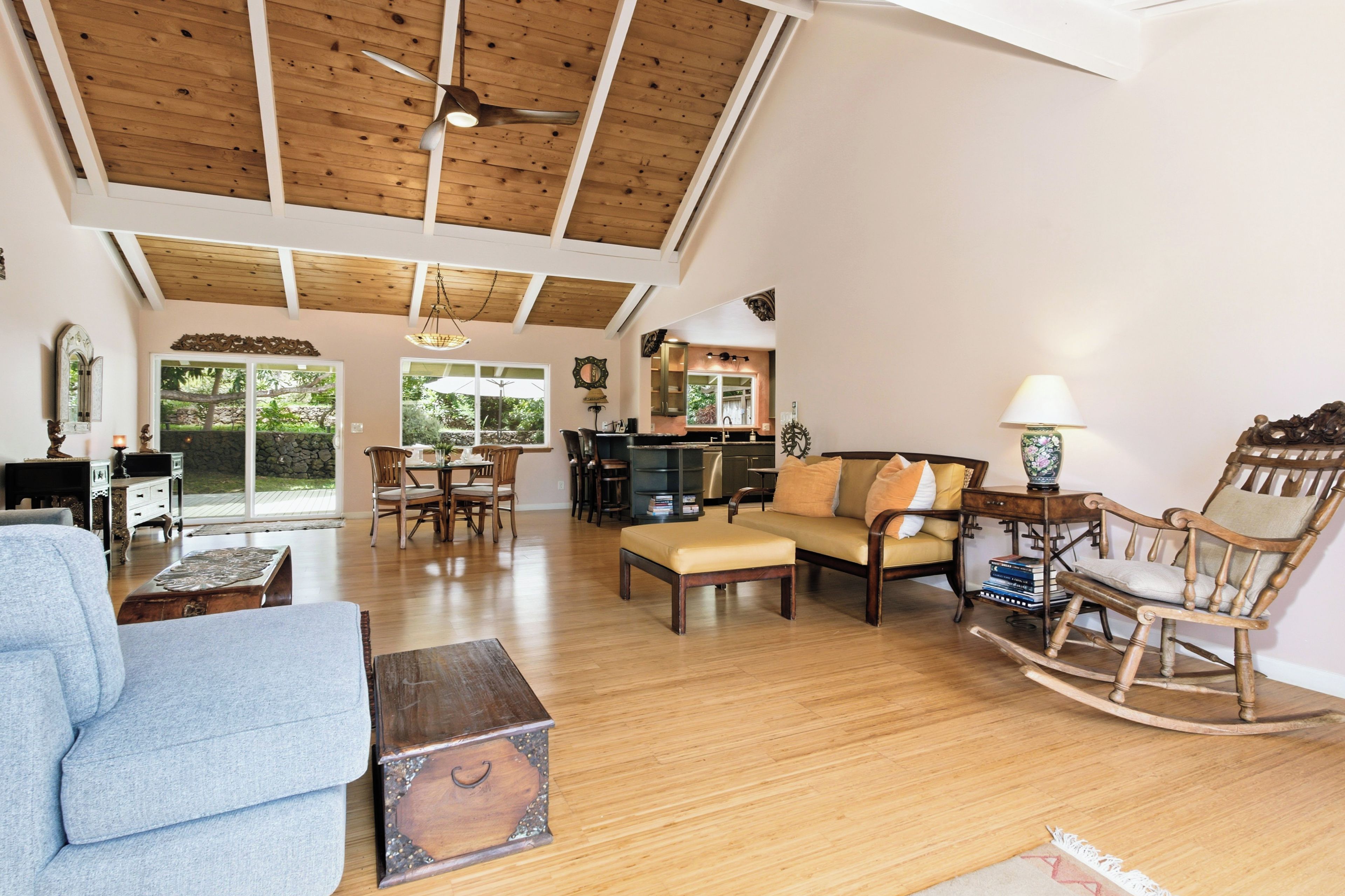 A spacious living room with wooden flooring, a high ceiling with exposed beams, a dining area with a table and chairs, and a kitchen in the background.