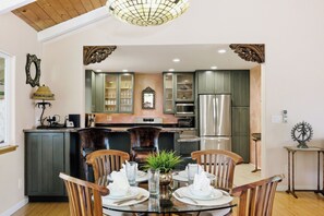 A modern kitchen with dark cabinets, a stainless steel refrigerator, and a dining area with a glass table and wooden chairs. - Lush Tropical Home Close to the Ocean - Mana Hale (Kihei)