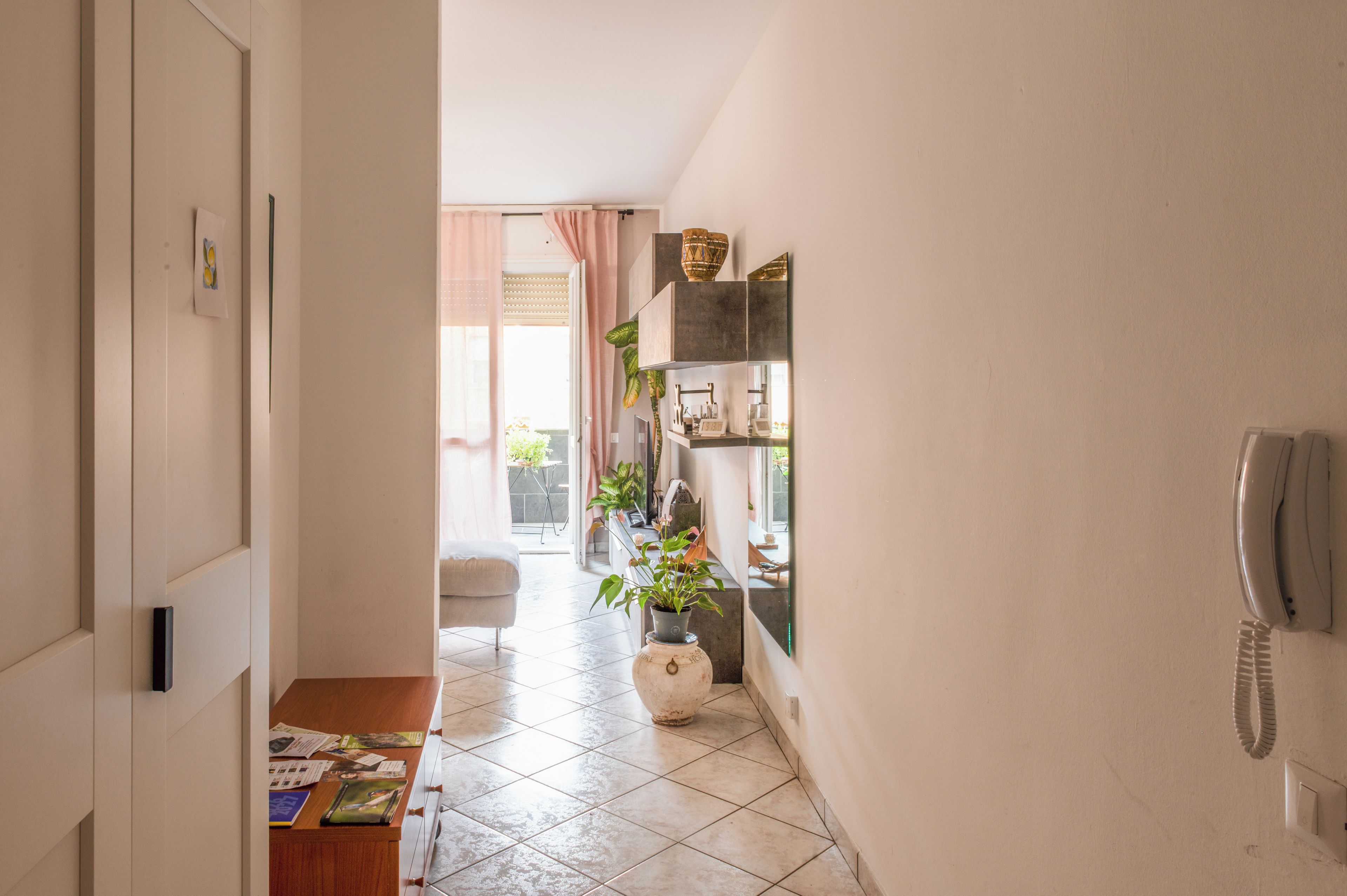 A hallway with a tiled floor, a wooden table with magazines, a potted plant, and a telephone on the wall.