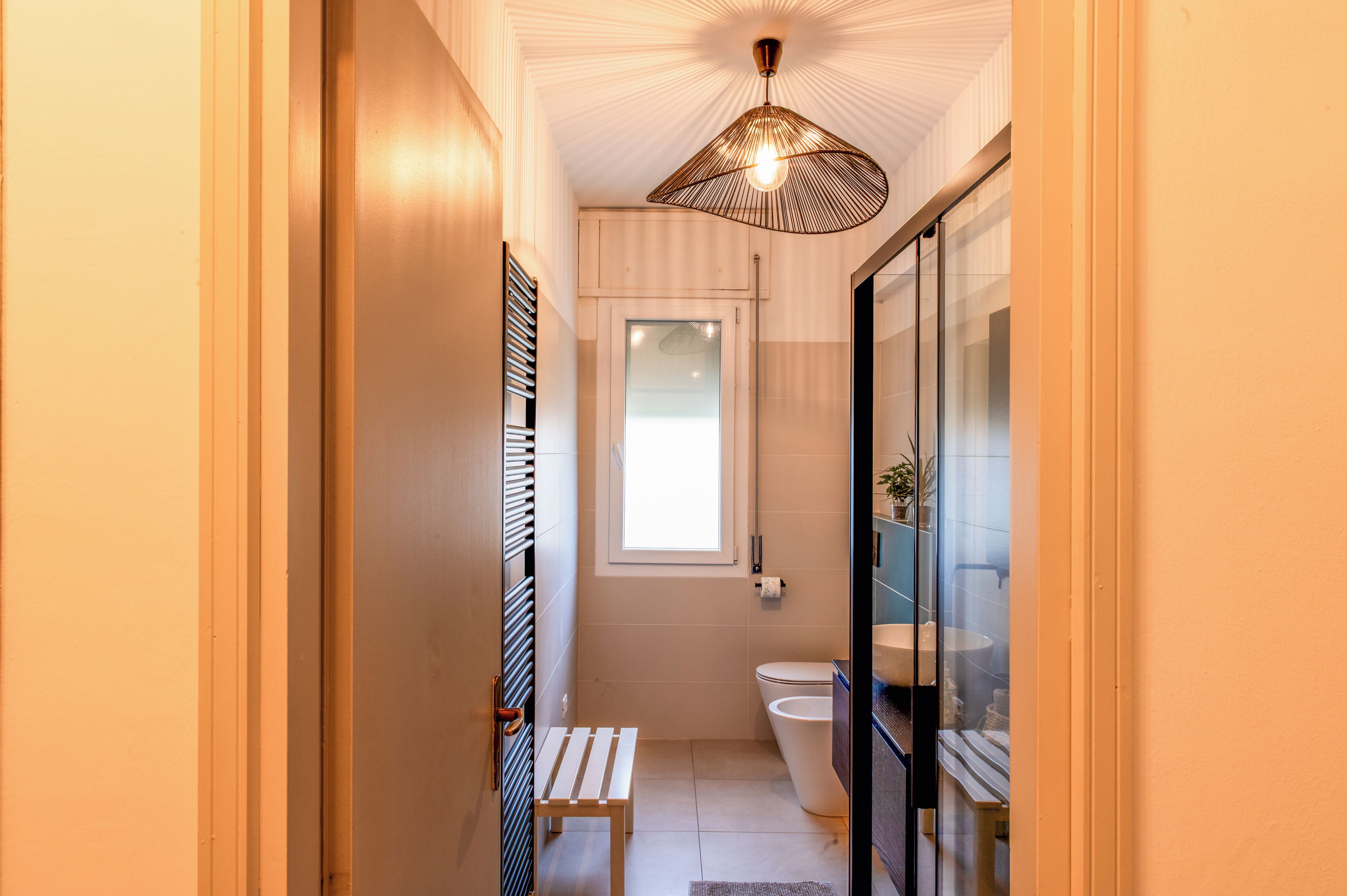 A narrow bathroom with a toilet, a bench, and a shower area.