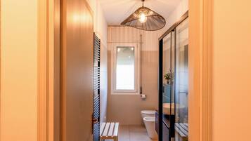 A narrow bathroom with a toilet, a bench, and a shower area.
