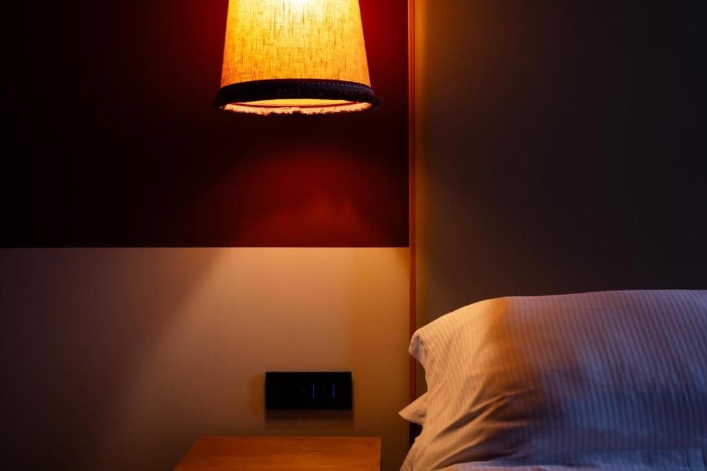 A dimly lit room with a bed, a wall-mounted light fixture, and a power outlet.