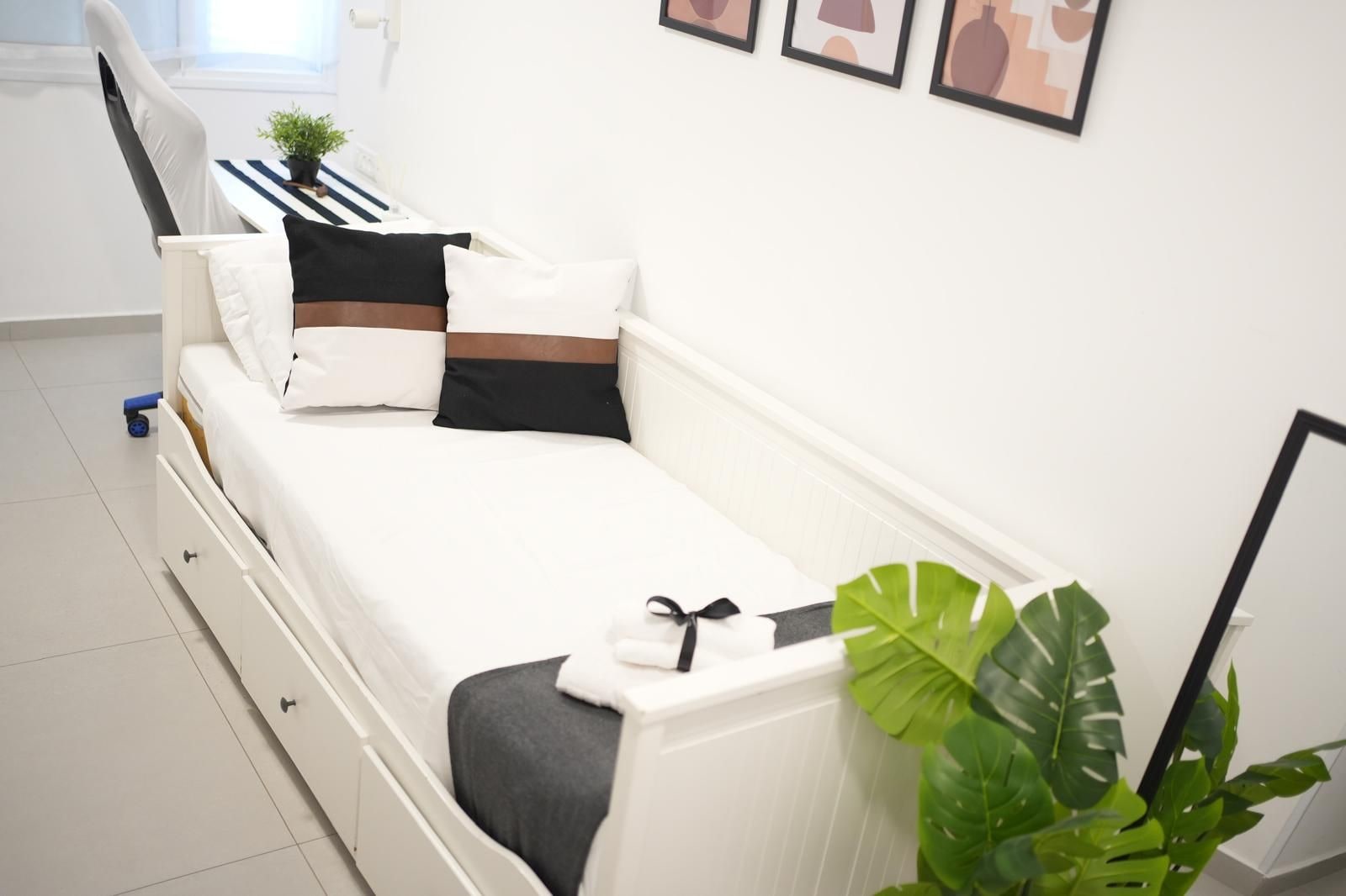 Una cama blanca con una manta gris, un almohadón negro y un almohadón marrón. Hay una toalla blanca sobre la cama. Una planta verde grande está en el suelo junto a la cama.