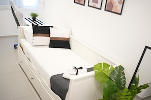 Een wit bed met een grijze deken, een zwart kussen en een bruin kussen. Er ligt een witte handdoek op het bed. Een grote groene plant staat op de vloer naast het bed.