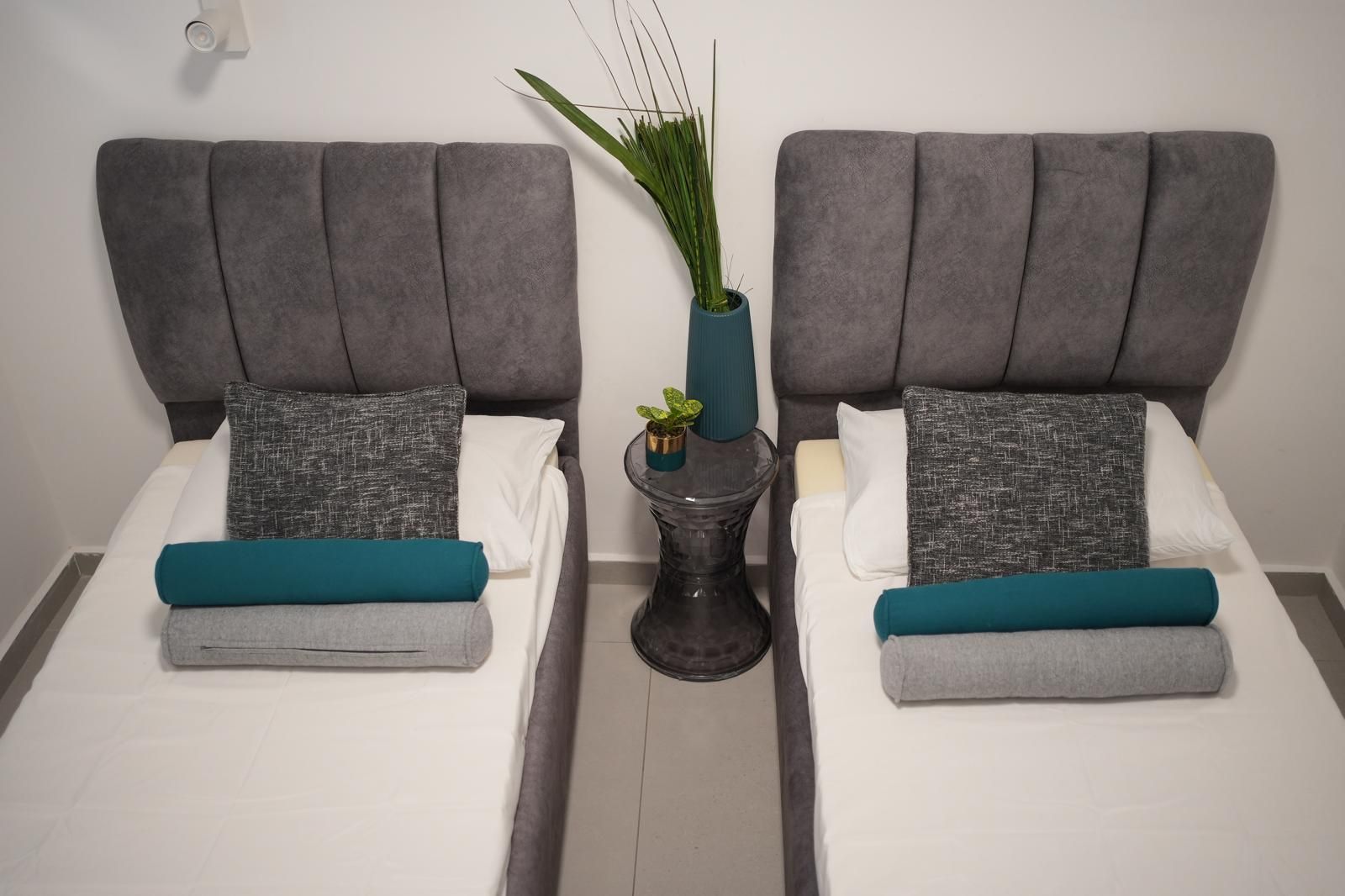 Une chambre d’hôtel avec deux lits, des têtes de lit grises, du linge de lit blanc, des coussins décoratifs aux tons turquoise et gris, et une petite plante en pot posée sur une table d’appoint.