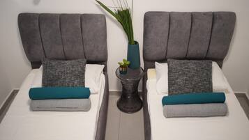 Une chambre d’hôtel avec deux lits, des têtes de lit grises, du linge de lit blanc, des coussins décoratifs aux tons turquoise et gris, et une petite plante en pot posée sur une table d’appoint.