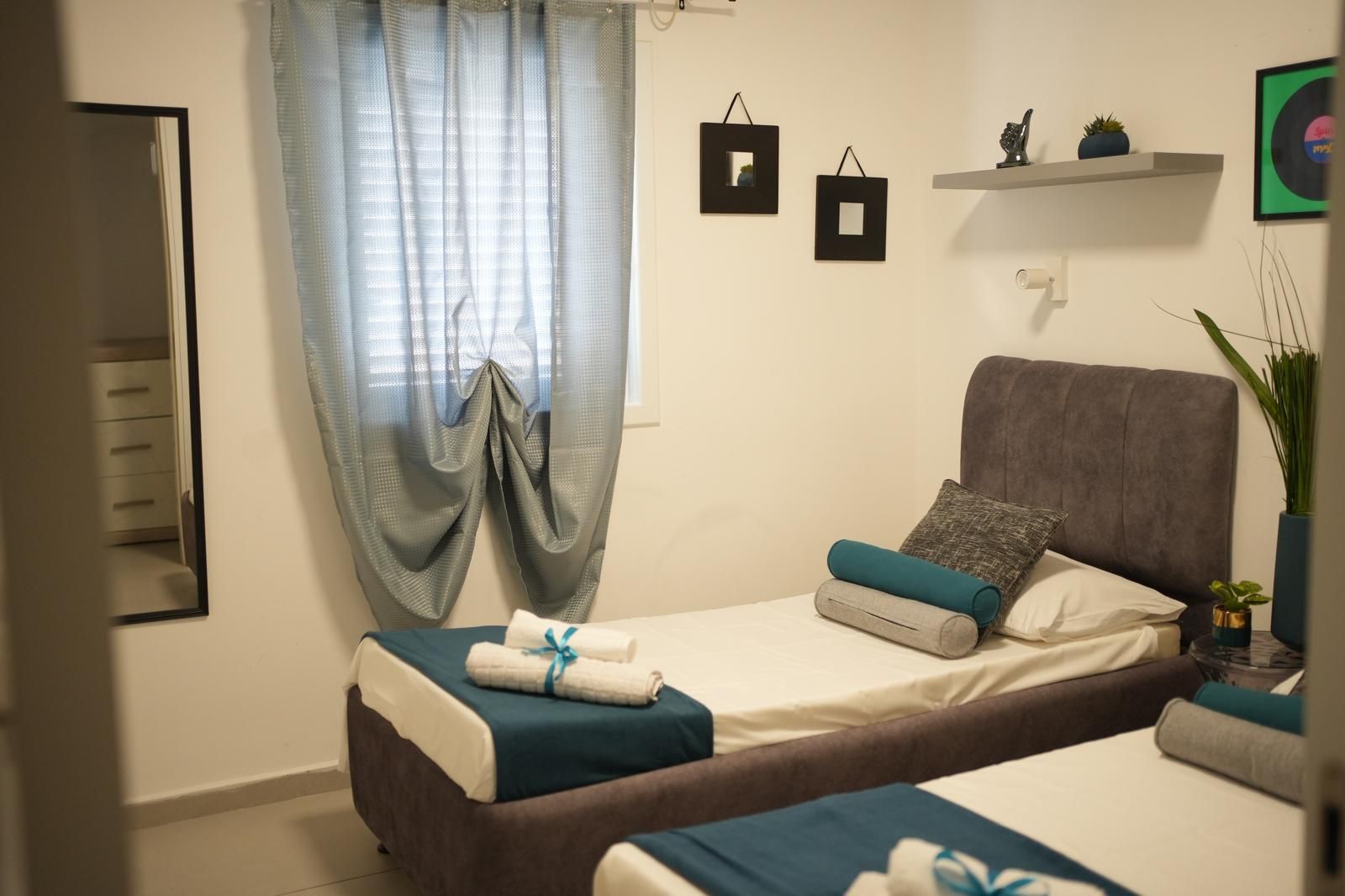 Una camera da letto con due letti, uno specchio, una finestra con tende, uno scaffale con oggetti decorativi e una pianta in vaso.