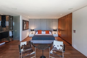 A modern hotel room with a large bed, two bedside tables with lamps, a glass shower enclosure, and a wooden wardrobe.